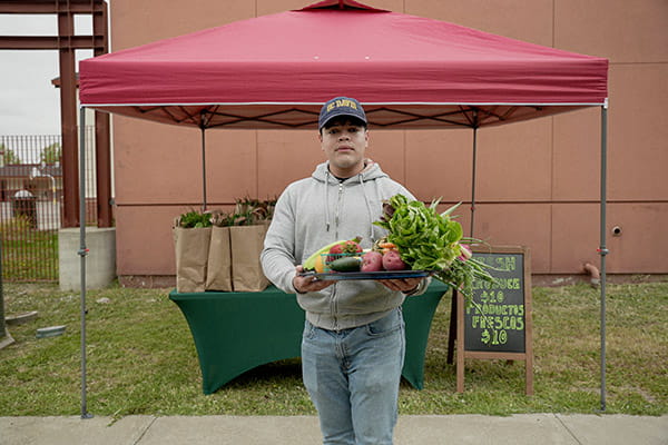 An Hispanic/Latino man is holding a large tray of freshly-picked produce in front of an outdoor Growing Together stand.