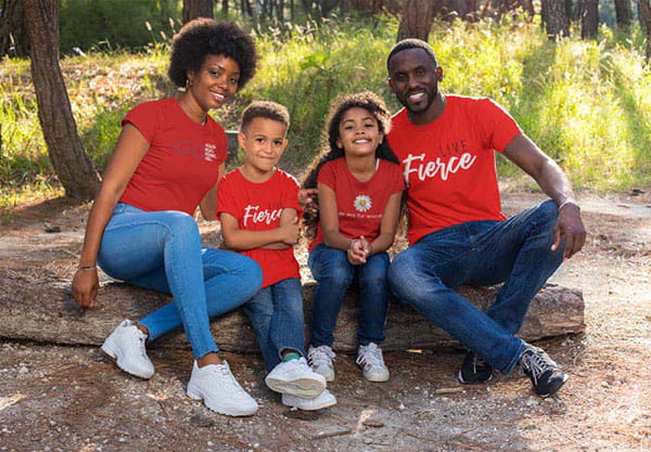 A family with two young children poses together sitting on the ground, all wearing red AHA t-shirts